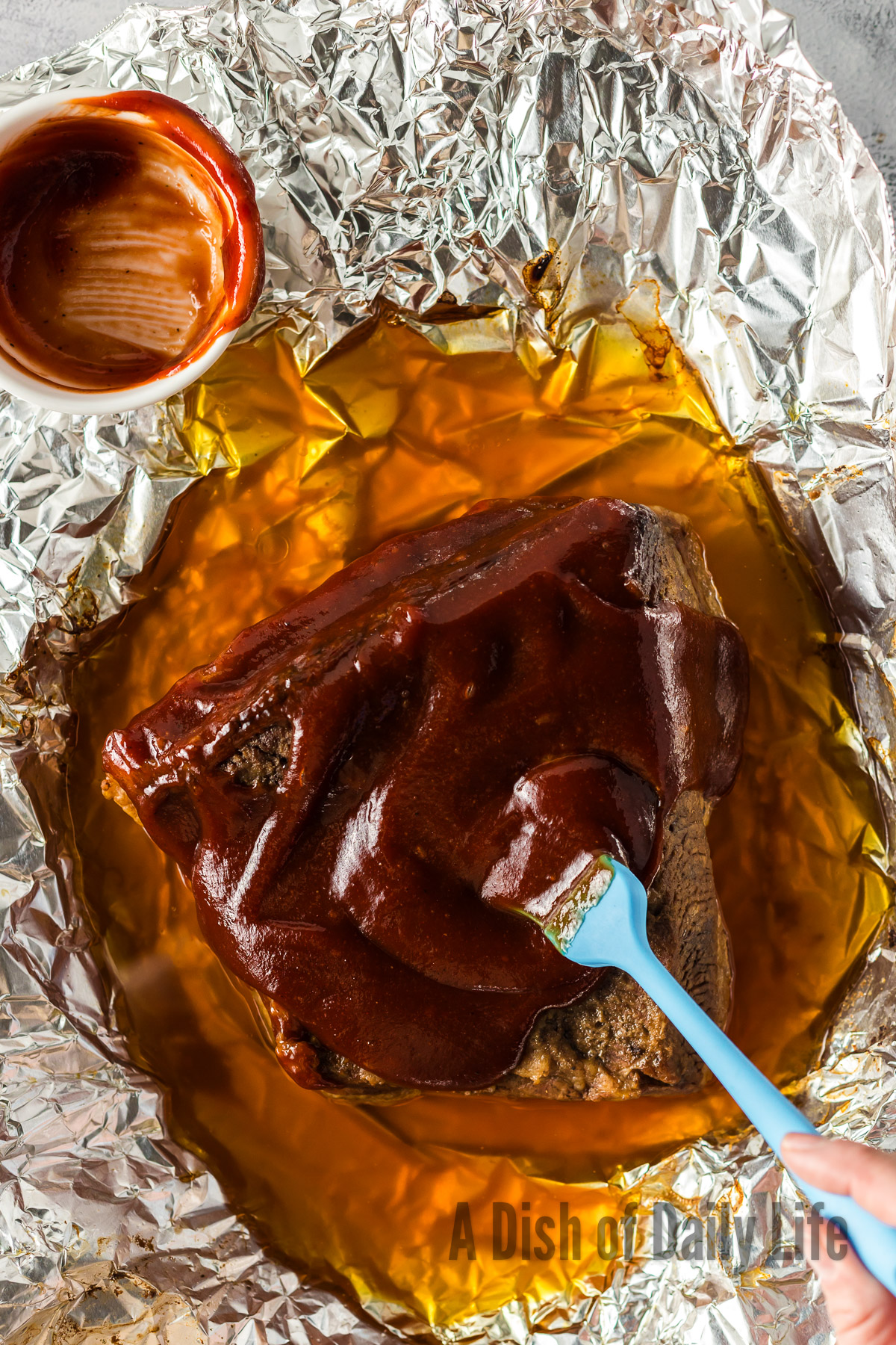 Baked brisket opened up with barbecue sauce being spread over it.