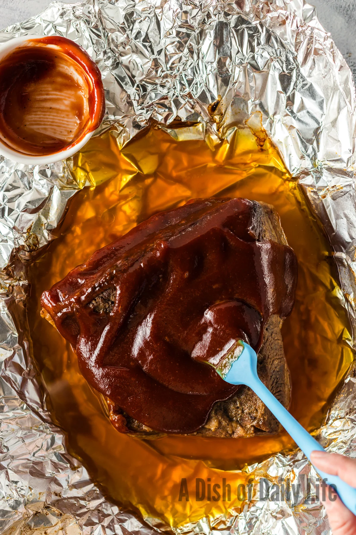 Baked brisket opened up with barbecue sauce being spread over it.