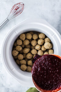 cranberry sauce being poured onto meatballs in crock pot.
