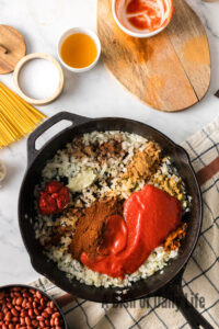 Onions, garlic, chili powder, cumin, cinnamon, allspice, ground cloves, cayenne, bay leaf, tomato sauce, tomato paste all added to a skillet.