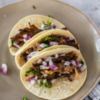 3 pork carnitas tacos on plate topped with red onion and cilantro.