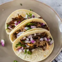 3 pork carnitas tacos on plate topped with red onion and cilantro.