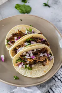 3 pork carnitas tacos on plate topped with red onion and cilantro.