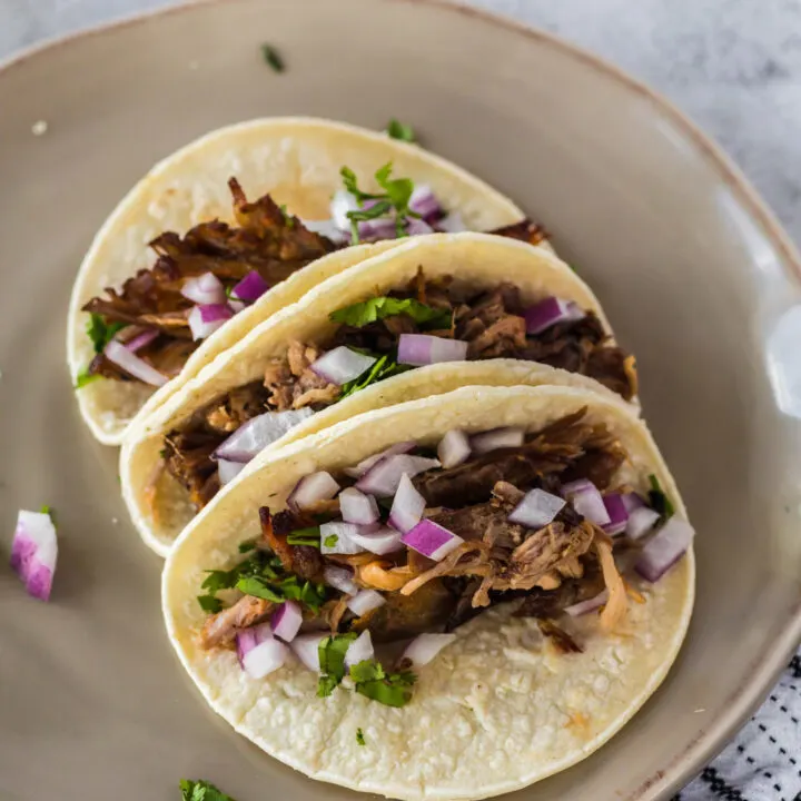 3 pork carnitas tacos on plate topped with red onion and cilantro.